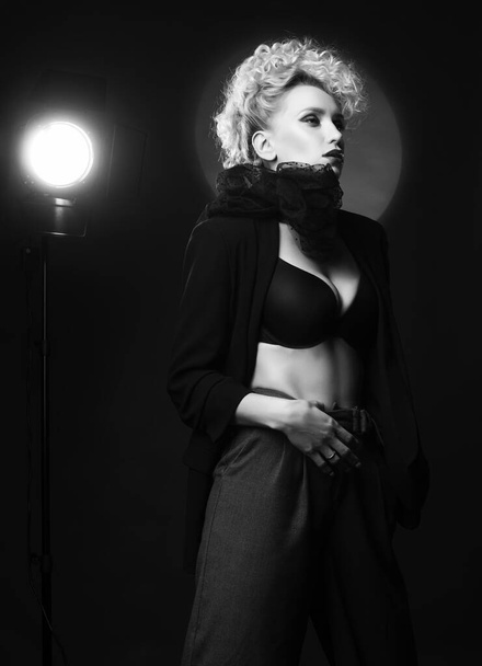A beautiful blonde girl with an elegant hairstyle and large breasts, wearing a bra, trousers, scarf and a blazer, fashinable poses near the spotlight. Trendy, monochrome, black and white, noir design - Zdjęcie, obraz