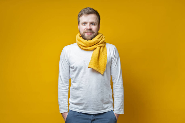 Attractive bearded man in a knitted scarf stands with hands folded in pockets and looks with a cute smile, on a yellow background. - Photo, Image