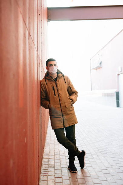 un jeune homme vêtu d'une veste brune avec un masque médical sur le visage dans la rue, près du mur rouge. protection contre le virus
 - Photo, image
