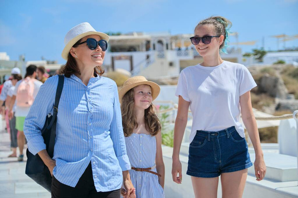 Mother walking with two daughters holding hand in famous tourist village Oia Santorini island. Happy woman and girls on sunny summer day walking together, family vacation - Foto, afbeelding