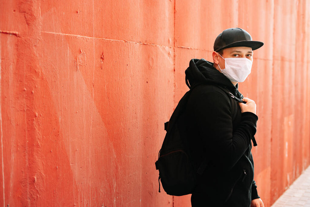 Een jongeman in zwarte sportkleding en een honkbalpet met een medisch masker op zijn gezicht bij de rode muur. bescherming tegen het virus - Foto, afbeelding