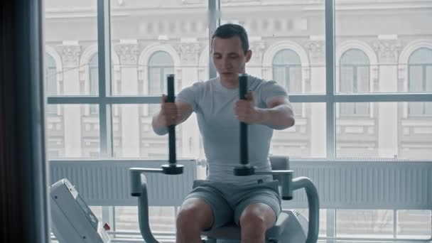 Modern spor salonu. Beyaz tişörtlü genç bir adam eğitim aletleri üzerinde kol egzersizi yapıyor. - Video, Çekim