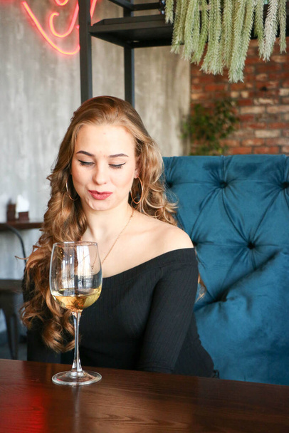Beautiful elegant blond girl wearing a black dress is relaxing in a cafe. Advertising,fashion,commercial design.Girl in a cafe in a black dress. Beautiful girl in a cafe. Bright red lips and nails. Curly long hair with a glass of wine - Photo, image