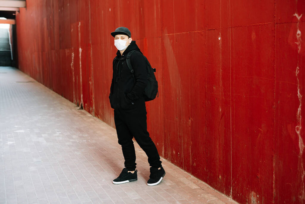 un jeune homme en tenue de sport noire et une casquette de baseball avec un masque médical sur le visage près du mur rouge. protection contre le virus
 - Photo, image