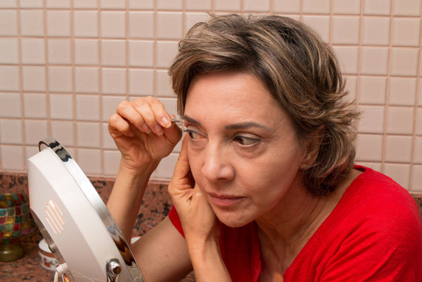 Женщина выщипывает брови пинцетом перед зажженным зеркалом
 - Фото, изображение