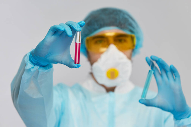 εστίαση στο ροζ υγρό του βιολογικού κινδύνου στο δοκιμαστικό σωλήνα στο βραχίονα του βιοχημικού φορώντας προστατευτική στολή με μπλε γάντια και κίτρινα γυαλιά - Φωτογραφία, εικόνα