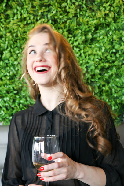 Beautiful elegant blond girl wearing a black dress is relaxing in a cafe. Advertising,fashion,commercial design.Girl in a cafe in a black dress. Beautiful girl in a cafe. Bright red lips and nails. Curly long hair with a glass of wine on a green back - Foto, imagen