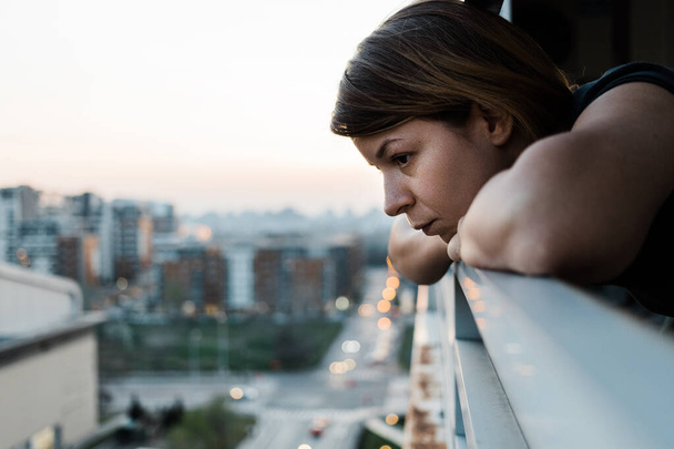 Junge traurige Frau blickt durch Balkon eines Mehrfamilienhauses nach draußen - Foto, Bild