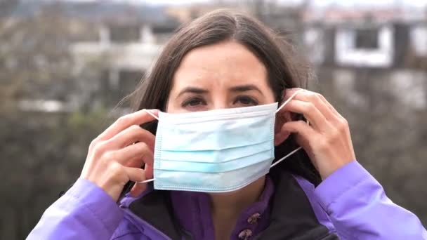 Young woman putting a protective surgical face mask during the Coronavirus disease (COVID-19) outbreak epidemic. Close up portrait with a single use blue protection mask on the face. Looking at camera - Materiał filmowy, wideo