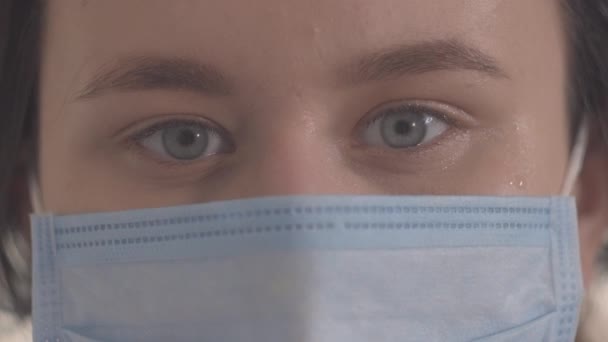 Extreme close-up of grey eyes of young woman in protective mask. Beautiful brunette Caucasian girl looking at camera. Quarantine measures, health care, isolation, safety. - Кадри, відео