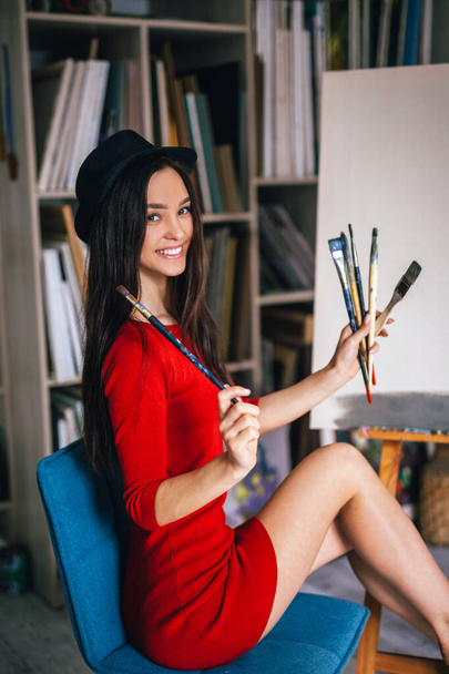 woman artist sits on a blue chair near an easel with brushes in her hands - 写真・画像