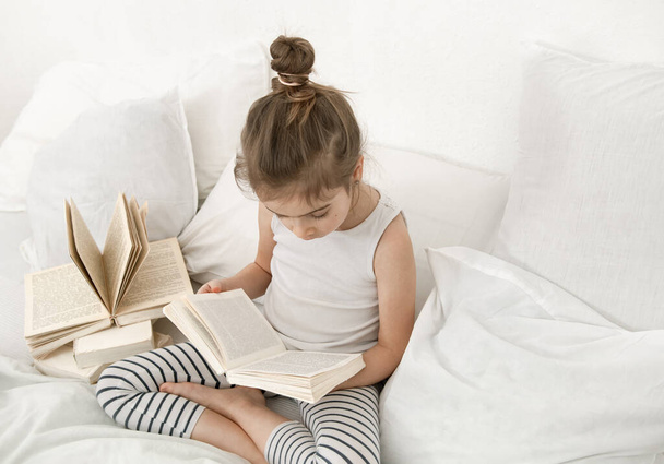 Cute little girl reading a book on the bed in the bedroom. The concept of education and family values . - Fotó, kép