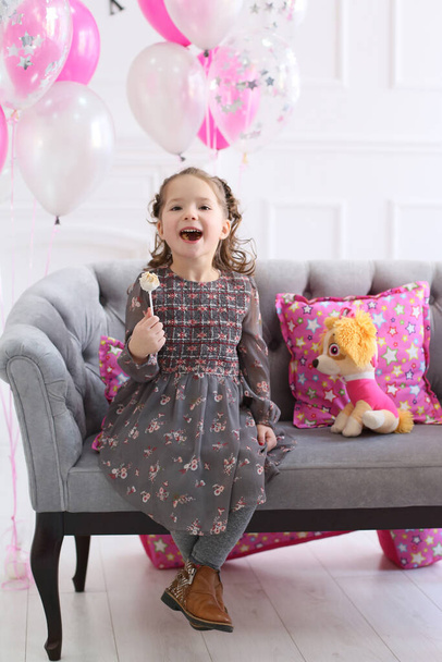pretty curly girl in a gray flower dress laughing sitting on a sofa with candy in hand - Photo, Image