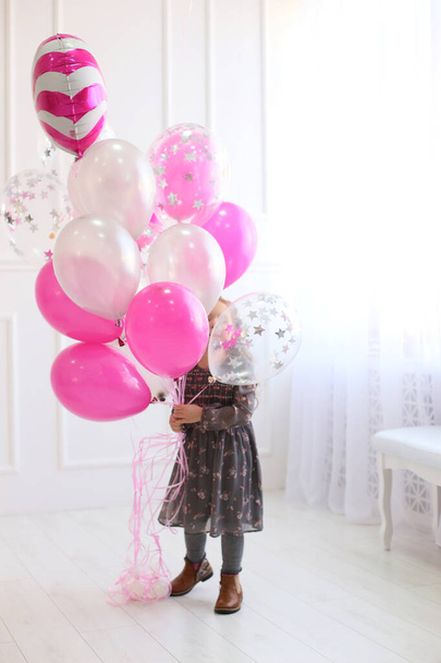 Gri çiçekli bir elbise ve kahverengi ayakkabılı küçük bir kız büyük bir demet gümüş, şeffaf ve parlak pembe balonlarla beyaz bir stüdyoda duruyor. - Fotoğraf, Görsel