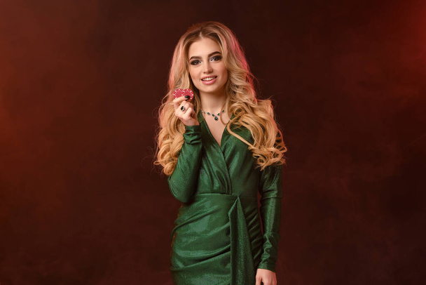 Blonde curly model, bright make-up, in green stylish dress and jewelry. Smiling, showing two red chips, posing on brown smoky background. Close-up - Foto, immagini