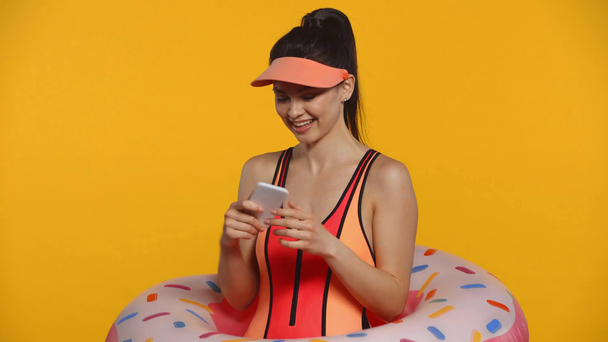 Uśmiechnięta dziewczyna w stroju kąpielowym i pierścień kąpielowy za pomocą smartfona izolowanego na żółto - Materiał filmowy, wideo
