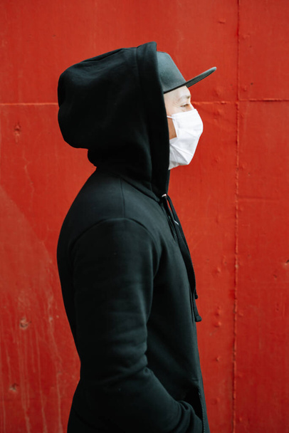 un giovane uomo in abbigliamento sportivo nero e un berretto da baseball con una maschera medica sul viso vicino al muro rosso. protezione dal virus
 - Foto, immagini