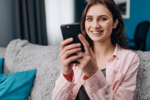 Cheerful girl sitting on couch and using smartphone - Фото, изображение