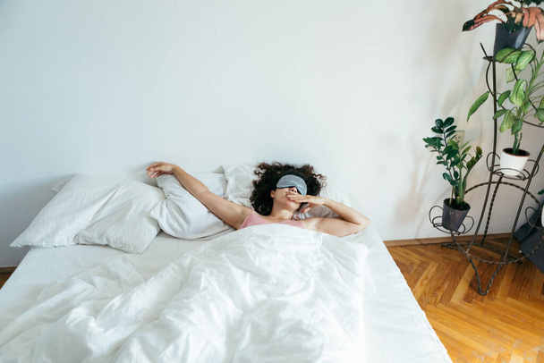 γυναίκα με μάσκα ύπνου στο κρεβάτι πρωί φως μέσα από κουρτίνες. λευκά φύλλα - Φωτογραφία, εικόνα
