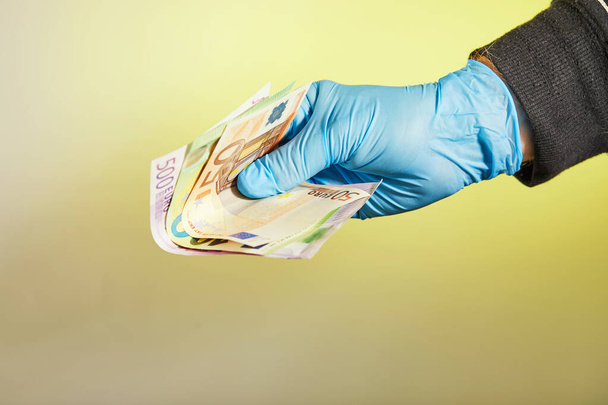La mano de un hombre con un guante médico azul sostiene el dinero en forma de billetes de euro sobre un fondo amarillo. Protección contra virus y bacterias. Concepto. Covid-19 o Coronavirus
. - Foto, Imagen