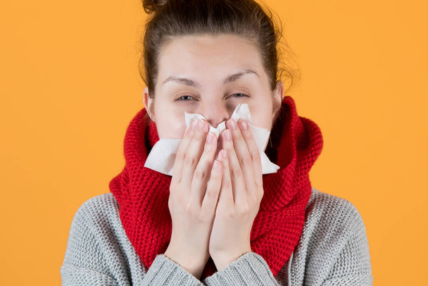 a sickly-looking girl in a scarf squints, sneezing into a scarf on yellow background - Fotoğraf, Görsel
