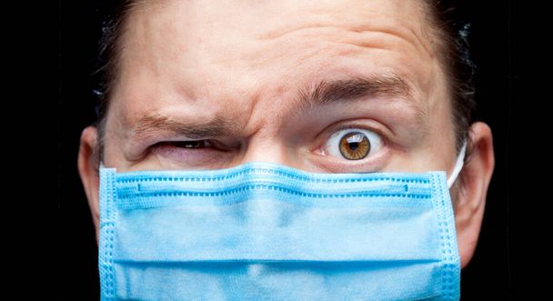 a man portrait in a medical mask with wide open surprised one eye and squinted second eye, close up of a face with suspiciously displeased emotion of the coronovirus covid 19 pandemic. - Fotografie, Obrázek