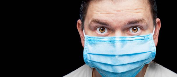 männliches Arztporträt in blauer medizinischer Maske mit einem überraschten Blick, der seine Augenbrauen hochzieht, Großaufnahme-Banner mit Kopierraum isoliert auf schwarzem Hintergrund für medizinisches Thema. - Foto, Bild