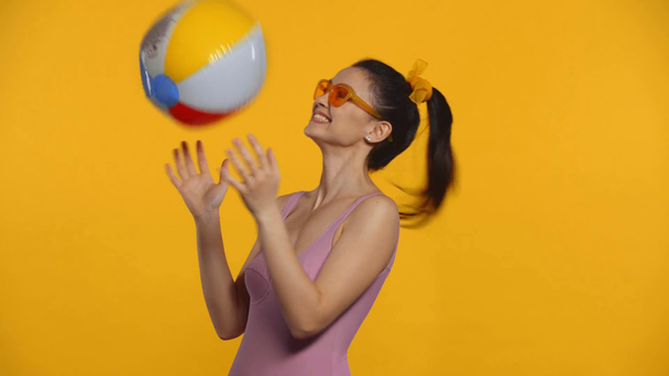 Chica sonriente en traje de baño lanzando bola inflable aislada en amarillo
 - Metraje, vídeo