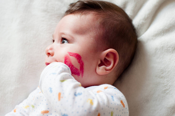 mignon nouveau-né gros plan couché dans le lit souriant avec une marque de rouge à lèvres baiser sur sa joue
. - Photo, image
