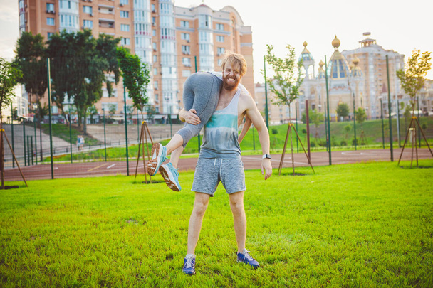 Een jong Kaukasisch echtpaar speelt en verheugt zich na de training op een grasveld in een zomerpark. Het thema van gezamenlijke opleiding. Gezonde atletische familie. jong sport paar gelukkig samen buiten. - Foto, afbeelding