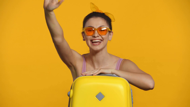 Pozitivní dívka v slunečních brýlích mává rukou v blízkosti kufru izolované na žluté - Záběry, video