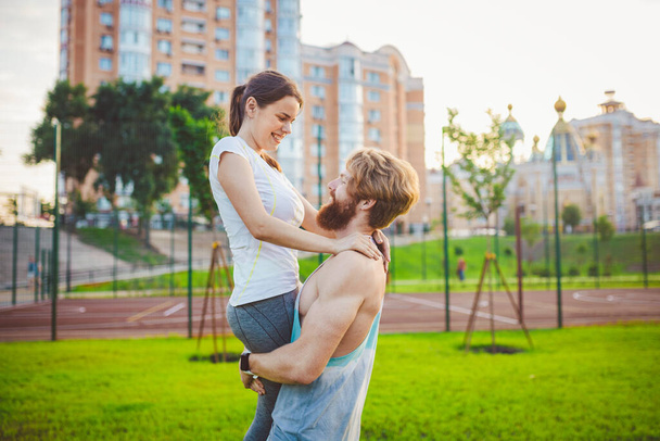 A young Caucasian couple plays and rejoices after training on a lawn in a summer park. The theme of joint training. Healthy athletic family. Young sport couple happy together outdoors. - Photo, Image
