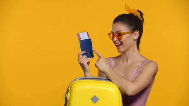 Χαμογελαστή γυναίκα που κατέχει διαβατήριο και αεροπορικό εισιτήριο κοντά σε βαλίτσα απομονωμένη στο κίτρινο - Πλάνα, βίντεο