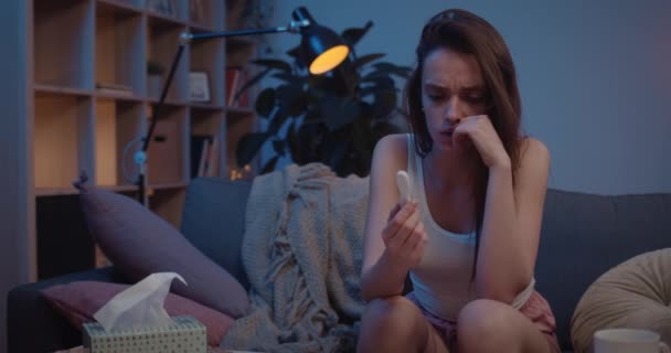 Anxious brunnette teenage girl holding pregnancy test while sitting on sofa.Front view of young upset woman looking worried about unwanted pregnancy. Home background. - Кадры, видео