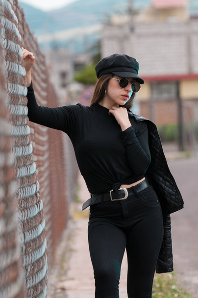 Retrato de mujer hermosa en gorra negra y camiseta negra y gafas de sol descansando en bastidores
 - Foto, Imagen