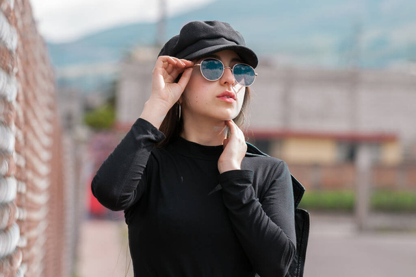 Portrait of beautiful woman in black cap and black t-shirt and sunglasses resting on racks - Foto, Bild