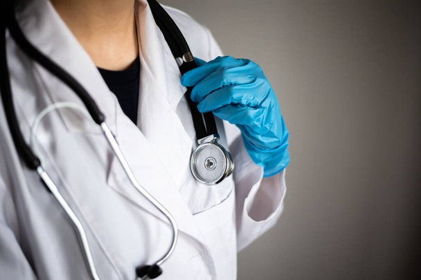 Close-up van een hand in een blauwe medische handschoen met stethoscoop in de nek van een arts. Ziekenhuiscontrole op coronavirus nCov2019, Doktersbezoek tijdens epidemische pandemie - Foto, afbeelding