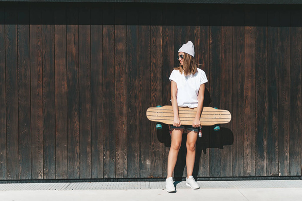 Attractive woman in sunglasses and hat standing at the wall with longboard - Fotoğraf, Görsel