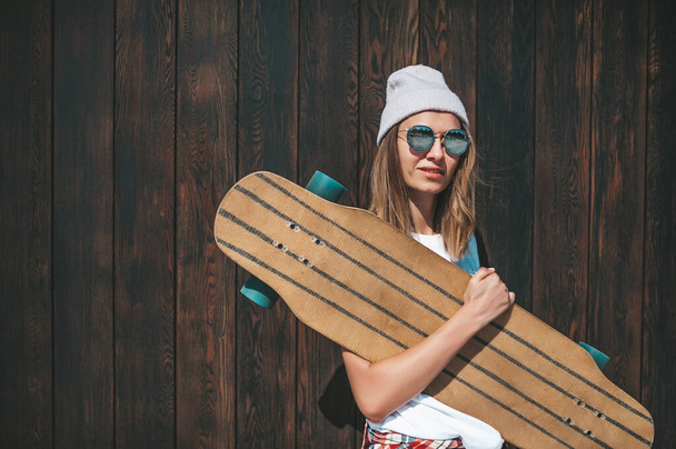 Attractive woman in sunglasses and hat standing at the wall with longboard - Photo, Image