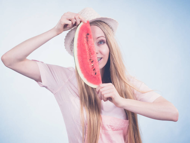 Happy cheerful teenage girl ready for summer wearing sun hat holding sweet fruit red juicy watermelon, on blue - Фото, изображение