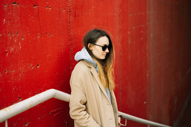Ein junges Mädchen in braunem Mantel und Brille steht auf der Straße neben einer roten Wand. Straßenstil - Foto, Bild