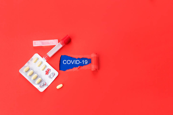 青色の背景に単語covid-19で赤い紙を回します。黄色の錠剤と血液フェンスのためのバイアル。コロナウイルス. - 写真・画像
