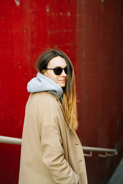 una joven con un abrigo marrón y gafas en la calle, de pie cerca de una pared roja. estilo de calle
 - Foto, Imagen