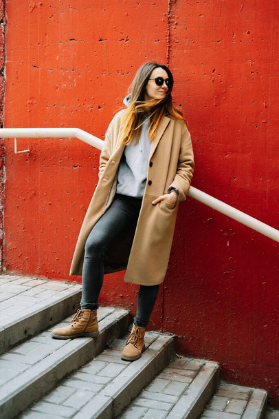 a young girl in a brown coat and glasses on the street, standing near a red wall. street style - Φωτογραφία, εικόνα