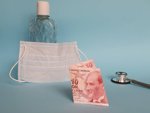 turkish banknote of ten lira, mask, bottle with gel alcohol and stethoscope on blue background - Fotó, kép