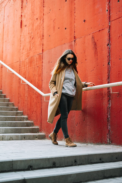 Caddede kırmızı duvarın yanında duran kahverengi ceketli ve gözlüklü genç bir kız. sokak biçimi - Fotoğraf, Görsel