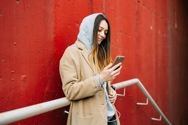 una joven con un abrigo marrón y gafas en la calle, de pie cerca de una pared roja utiliza su teléfono inteligente
 . - Foto, imagen