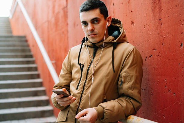 Caddede kırmızı duvarın yanında duran kahverengi ceketli genç bir adam akıllı telefonunu kullanıyor. . - Fotoğraf, Görsel