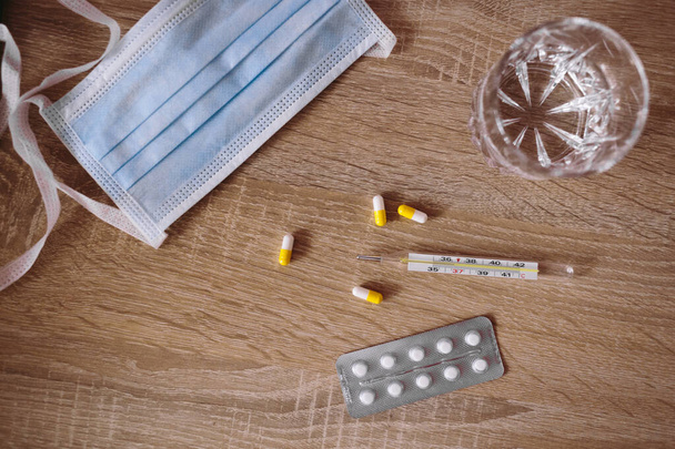 Medical mask, pills, thermometer and a glass of water on the wooden table. - Photo, image