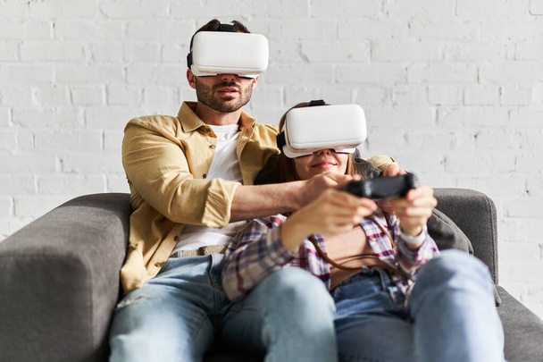 Junges lächelndes Paar mit VR-Brille spielt Videospiel mit Joystick und sitzt auf der Couch - Foto, Bild